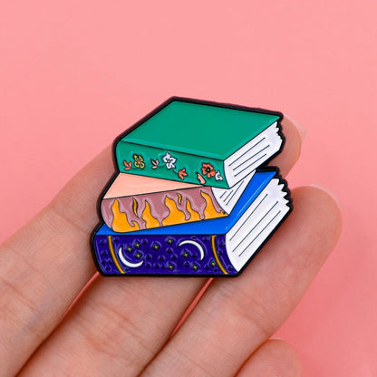 A hand displays an enamel pin with a stack of three books—green with flowers, orange with flames, and dark blue with stars and a moon—perfect for Booktroverts. The scene is set against a pink background.
