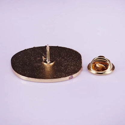 A My Cutesy Era gold enamel pin badge with a textured, round face lies flat on a light surface, its pointy pin and separate gold butterfly clutch backing displayed beside it.