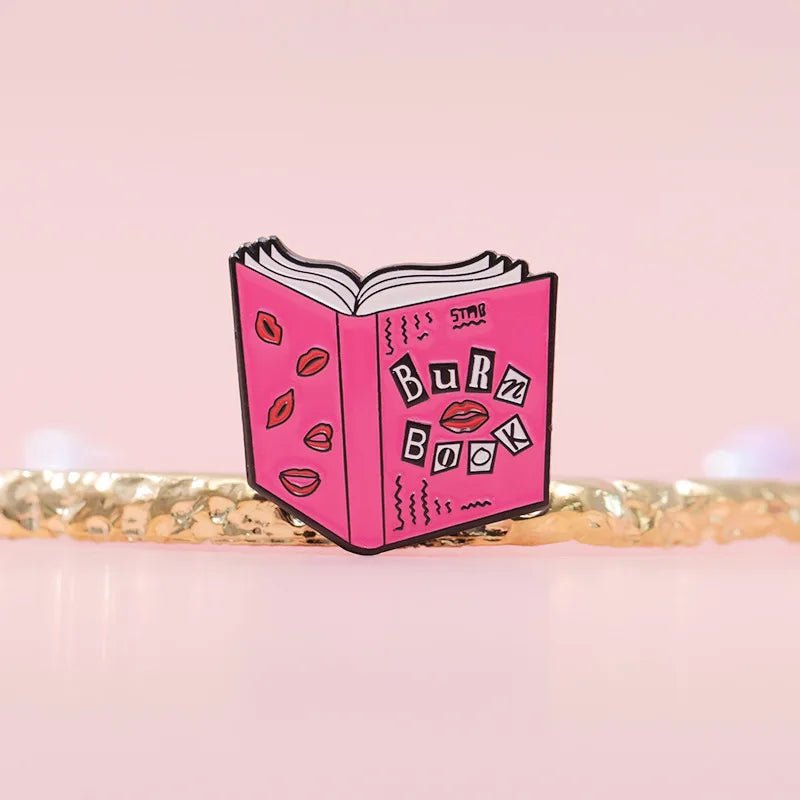 A My Cutesy Era pink enamel pin shaped like an open book labeled Burn Book with lip doodles—perfect for Mean Girls fans—shown on a soft pink background.