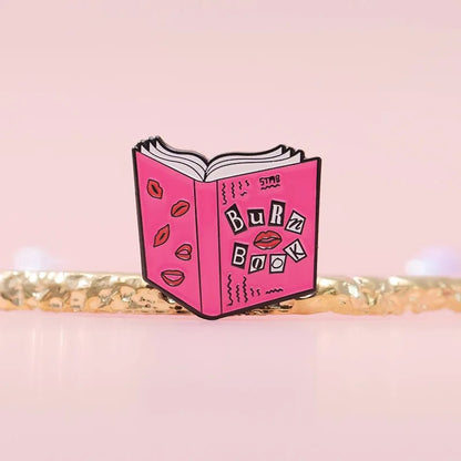 A My Cutesy Era pink enamel pin shaped like an open book labeled Burn Book with lip doodles—perfect for Mean Girls fans—shown on a soft pink background.