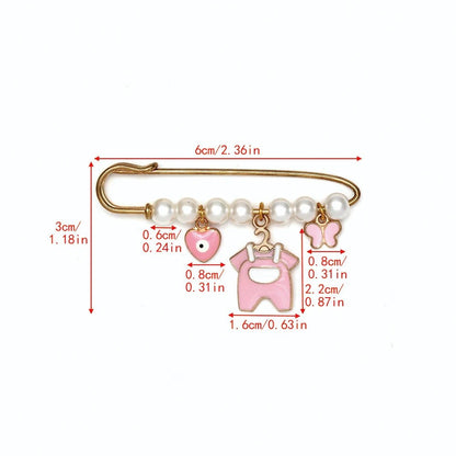 The Kiss Me Later gold safety pin features six pearls and three pink charms—a heart, overalls, and butterfly—creating a charming brooch. Red measurement lines indicate size in cm and inches; perfect as a baby shower gift.