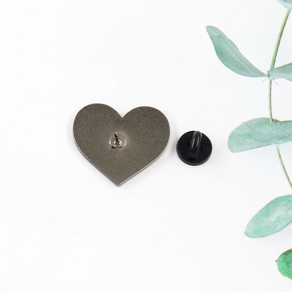 A Kiss Me Later pink enamel love heart pin with its black round backing separated, resting on a white surface with green leaves partly visible in the corner.