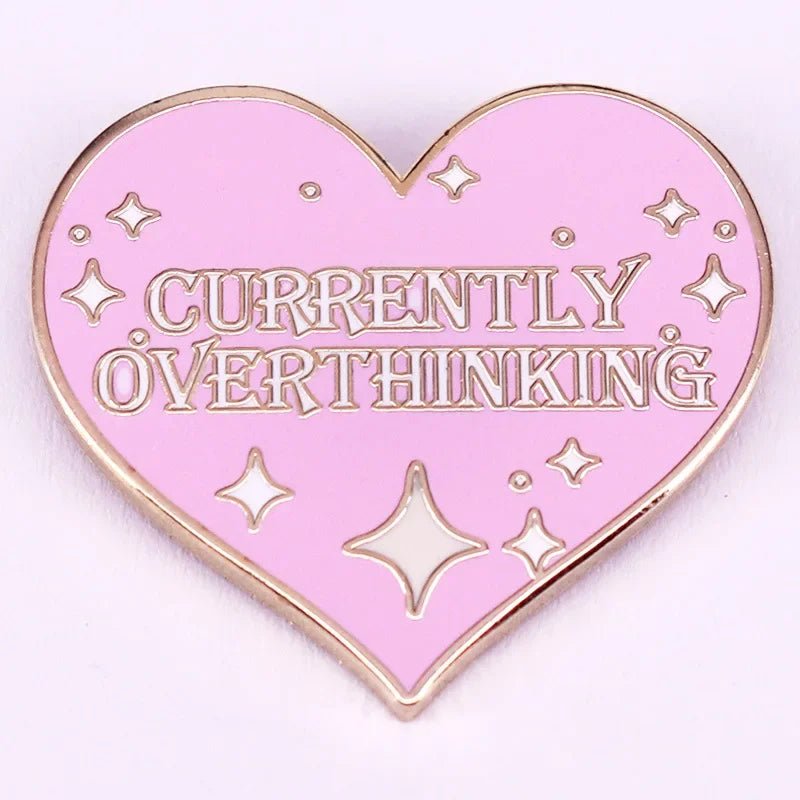 Sparkly enamel pin is a pink heart-shaped accessory featuring CURRENTLY OVERTHINKING in bold letters, accented with small star-like sparkles.