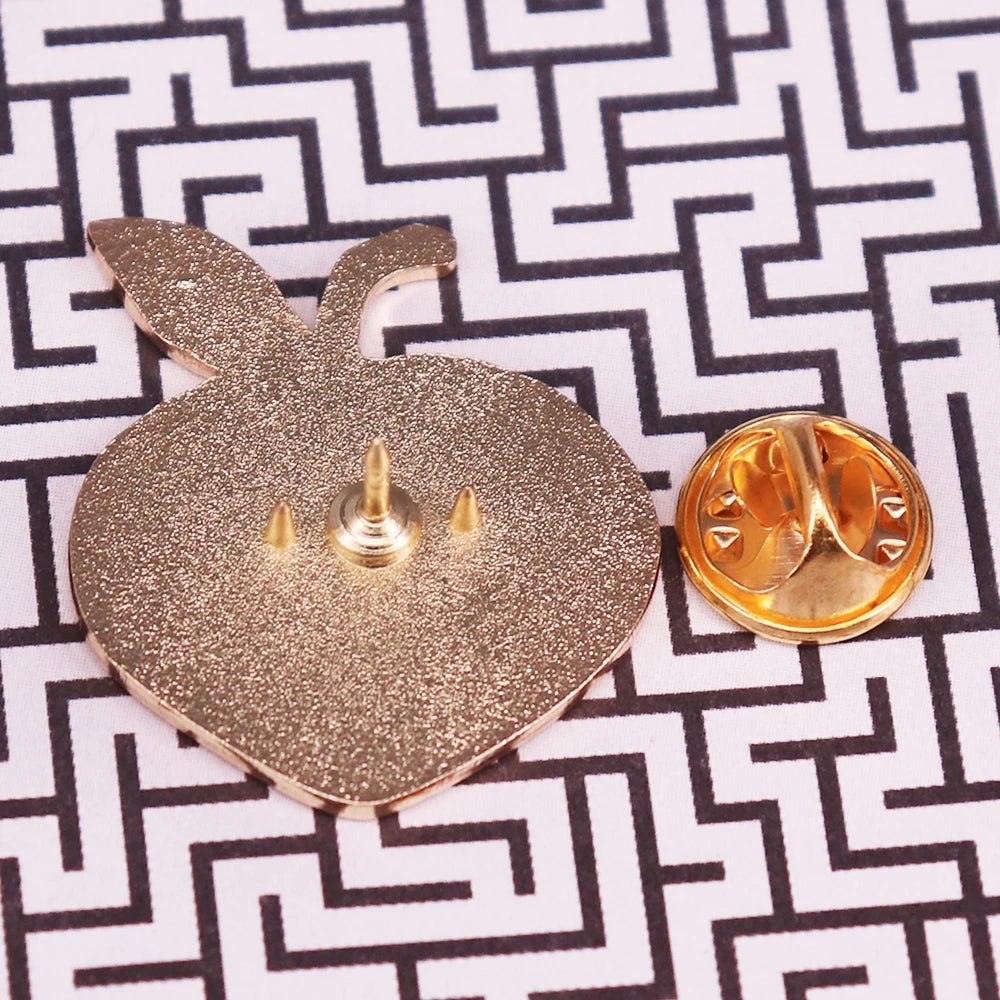 A gold-colored enamel apple pin shown from the back with its butterfly clutch off, rests on a black-and-white geometric surface—ideal for pairing with your favorite Bruise Me enamel pin.