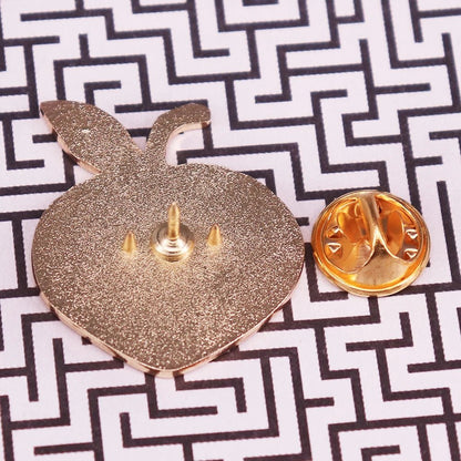 A gold-colored enamel apple pin shown from the back with its butterfly clutch off, rests on a black-and-white geometric surface—ideal for pairing with your favorite Bruise Me enamel pin.