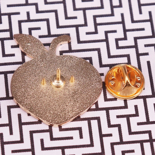 A gold-colored enamel apple pin shown from the back with its butterfly clutch off, rests on a black-and-white geometric surface—ideal for pairing with your favorite Bruise Me enamel pin.