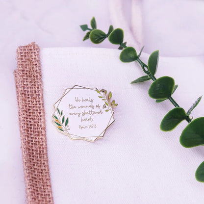 Enamel pin with a leafy border rests on white fabric beside a green vine. It reads, “He heals the wounds of every shattered heart. Psalm 147:3”—a touching symbol of comfort and hope.