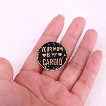 A hand holds a Kiss Me Later black and gold enamel pin with gold text, YOUR MOM IS MY CARDIO, accented by small hearts and star-like shapes.