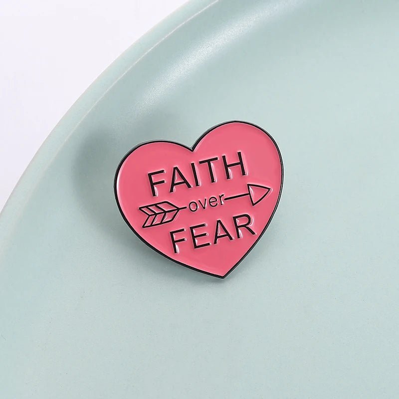 Love heart enamel pin, featuring Faith Over Fear and an arrow, is displayed on a light blue surface.