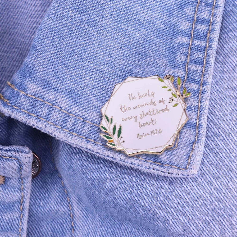 A gold and white floral enamel pin reading He heals the wounds of every shattered heart. Psalm 147:3, attached to a blue denim jacket—a beautiful reminder of hope and healing.