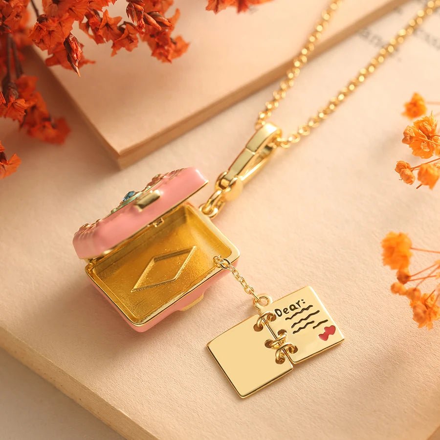 Gold pink enamel pendant necklace lies open, revealing a book pendant engraved with Dear: and a heart, surrounded by dried orange flowers and soft beige papers in the background.