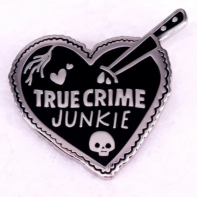 Enamel pin features a pink love heart with TRUE CRIME JUNKIE, a knife through the heart, a small skull, and blood drops, all set against a bold black and silver background.