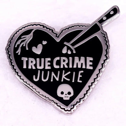 Enamel pin features a pink love heart with TRUE CRIME JUNKIE, a knife through the heart, a small skull, and blood drops, all set against a bold black and silver background.