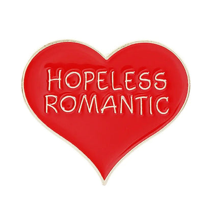 Heart-shaped enamel pin features red and gold colors, gold edges, and bold gold uppercase “HOPELESS ROMANTIC” lettering at the center.