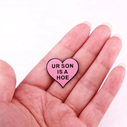 A hand holds a 1-inch enamel pin shaped like a pink heart with black text reading UR SON IS A HOE against a plain light background.