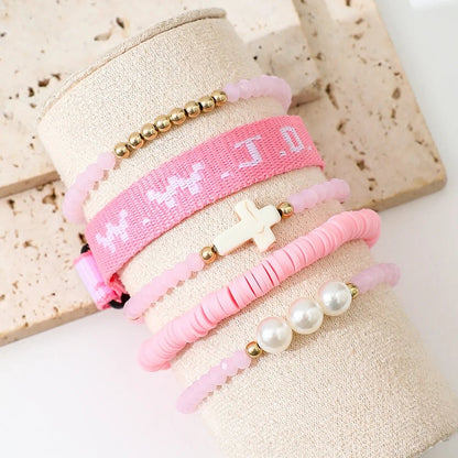 Kiss Me Later offers a set of four pink and gold beaded bracelets on fabric display, including a pink bracelet stack with woven WWJD, a white cross beaded piece, one with pearls, and one featuring small gold and pink beads.