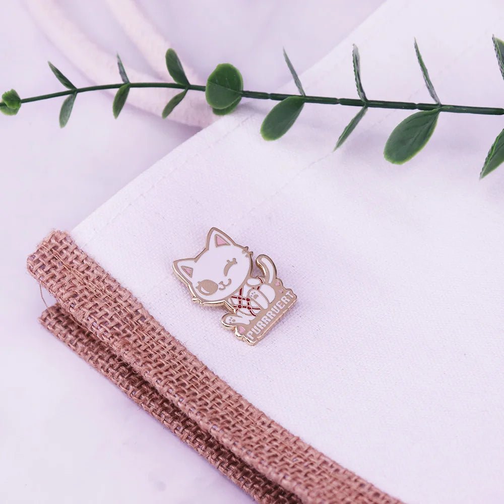 Waving cat pin with PURRRENDER is attached to a white bag with burlap trim, next to a green leafy sprig.