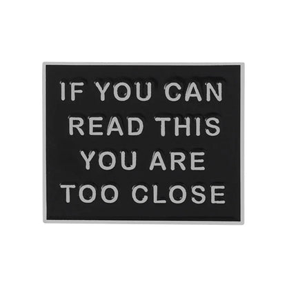 Black square pin features raised white letters saying IF YOU CAN READ THIS YOU ARE TOO CLOSE—a humorous accessory to add a fun touch to any outfit.