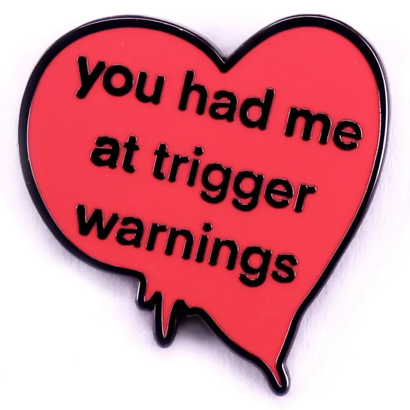 This red enamel pin is heart-shaped in pink with a dripping edge and features black text: you had me at trigger warnings.