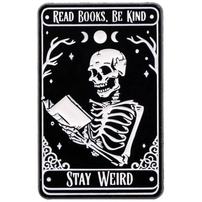 A Kiss Me Later pin features a black & white skeleton reading a book, surrounded by stars, branches, moons—evoking spooky vibes. Text above: Read Books, Be Kind; below: Stay Weird. Ideal for fans of unique skeleton hand accessories.