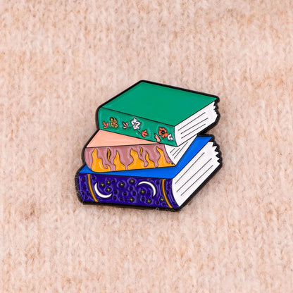 enamel pin features a colorful stack of three books—green with flowers, pink with flames, and blue with stars and moons—ideal for any book lover, shown on a soft beige textured fabric background.
