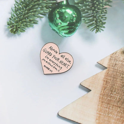 A Kiss Me Later wooden heart ornament with “Above all else, GUARD YOUR HEART, for everything you do flows from it. Proverbs 4:23” rests on a white surface next to a pink enamel pin and a wooden Christmas tree.