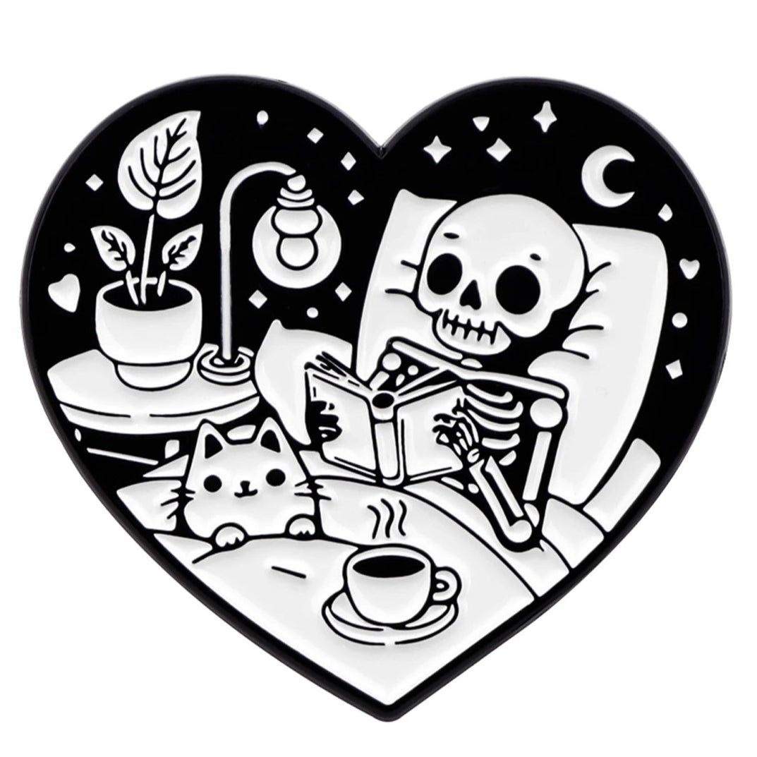 Kiss Me Later: A heart-shaped black and white print gives spooky vibes—thick-thighed skeleton reading in bed with a cat, tea, lamp, potted plant, and a crescent moon with stars glowing in the background.
