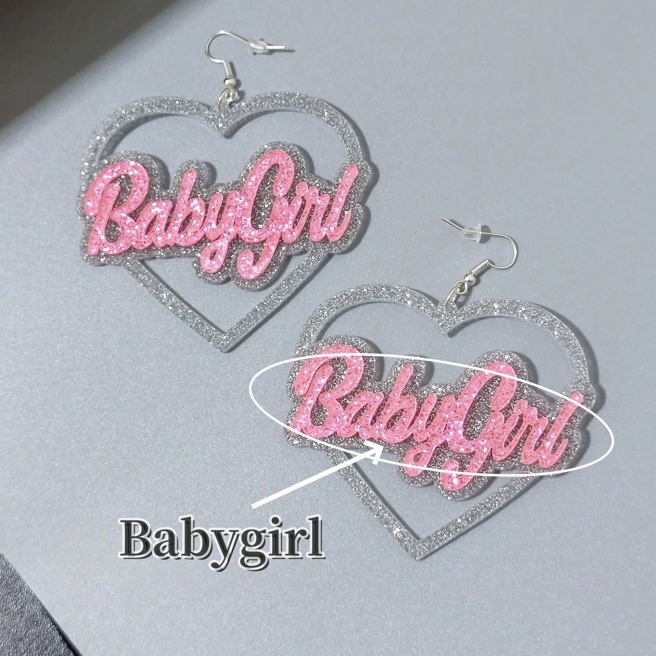 Two Kiss Me Later silver and pink glitter heart-shaped dangly earrings feature Babygirl in sparkly pink script. One earring lies flat, the other stands upright on a gray surface.