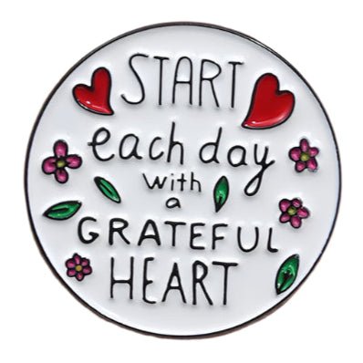 The My Cutesy Era enamel pin is round with red hearts, pink flowers, green leaves, and “Start each day with a grateful heart” in playful black lettering on a white background.