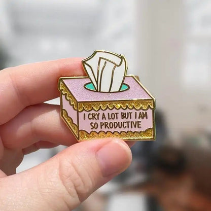 A hand holds a pink and gold enamel pin shaped like a tissue box with tissues popping out and the text “I cry a lot but I am so productive.” The softly blurred background adds focus to the pin.