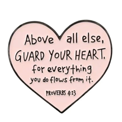 The Kiss Me Later pink love heart enamel pin reads: “Above all else, guard your heart, for everything you do flows from it. Proverbs 4:23.”.