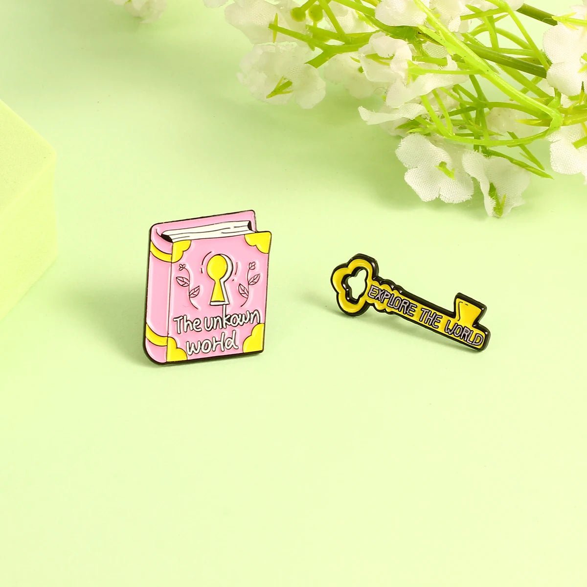 Enamel pins on a light green surface: a pink book labeled “The Unknown World” and a yellow key reading “Explore the World,” with white flowers in the background.