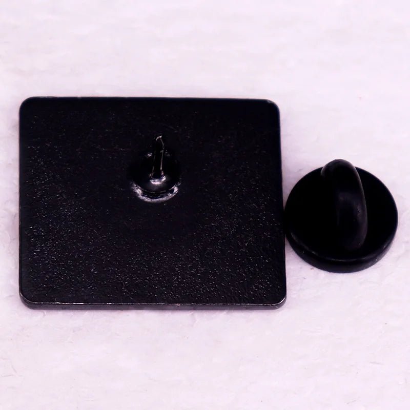 A square black enamel pin by Kiss Me Later is shown face down, displaying a central metal pin post with a separate round black rubber back—perfect for any book lover’s pin collection.
