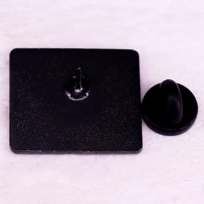 A square black enamel pin by Kiss Me Later is shown face down, displaying a central metal pin post with a separate round black rubber back—perfect for any book lover’s pin collection.