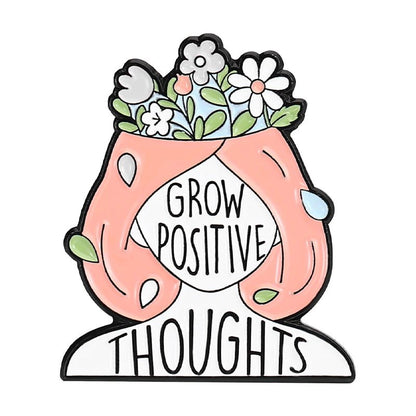 An illustration features a pink-haired person with their head open, blooming flowers inside, and Grow Positive Thoughts across the face—perfect inspiration for your next enamel pin design.