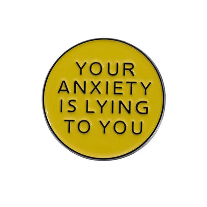 A limited edition round yellow enamel pin from Kiss Me Later features black text, YOUR ANXIETY IS LYING TO YOU, making it a motivational accessory for daily encouragement.