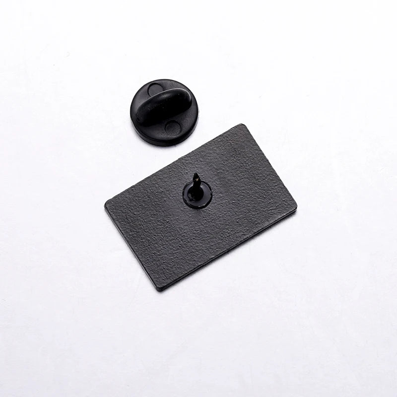 A rectangular black enamel pin lies face down on a white surface, revealing its single pointed pin post and a separate black metal butterfly clutch. This minimalist accessory is perfect for any feminist pin collection.