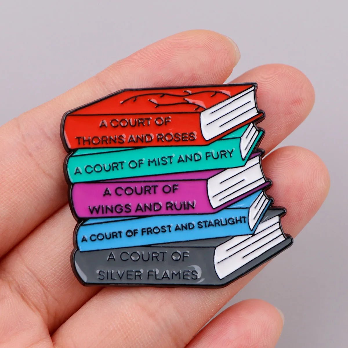 A hand holds a pin shaped like a stack of five colorful books, each labeled with titles from the A Court of Thorns and Roses series—an ideal accessory for any Booktrovert or library-loving fantasy fan.