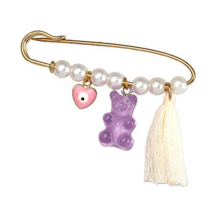 The Kiss Me Later gold safety pin features white beads, a pink evil eye heart charm, a purple gummy bear charm, and a cream tassel—ideal as a unique brooch or baby shower gift.