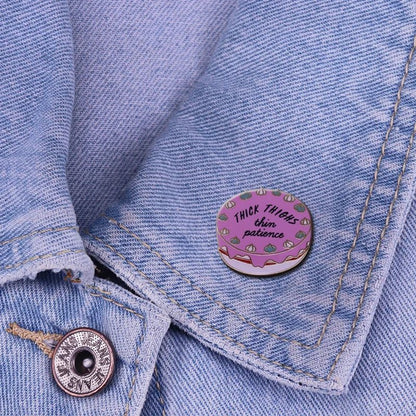 A round My Cutesy Era enamel pin reading Thick Thighs Thin Patience sits on a light blue denim jacket, featuring a pink background with playful green and white doodles.