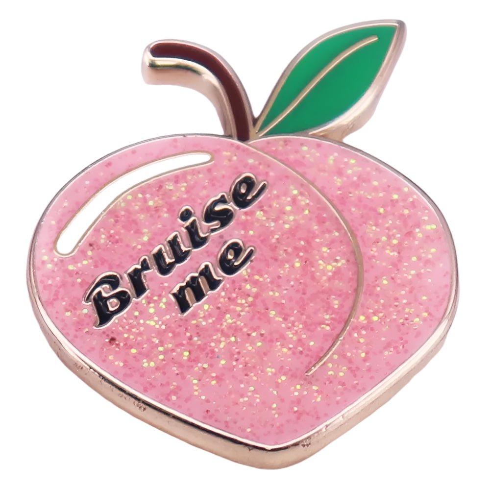 The My Cutesy Era pink glitter peach enamel pin features a peach shape with a green leaf and Bruise Me in black lettering across the front.