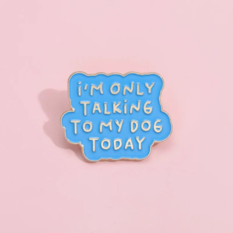 A blue enamel pin shaped like a speech bubble with IM ONLY TALKING TO MY DOG TODAY in yellow, is shown on a light pink background.