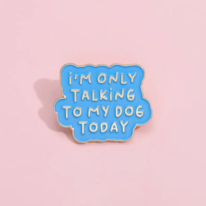 A blue enamel pin shaped like a speech bubble with IM ONLY TALKING TO MY DOG TODAY in yellow, is shown on a light pink background.