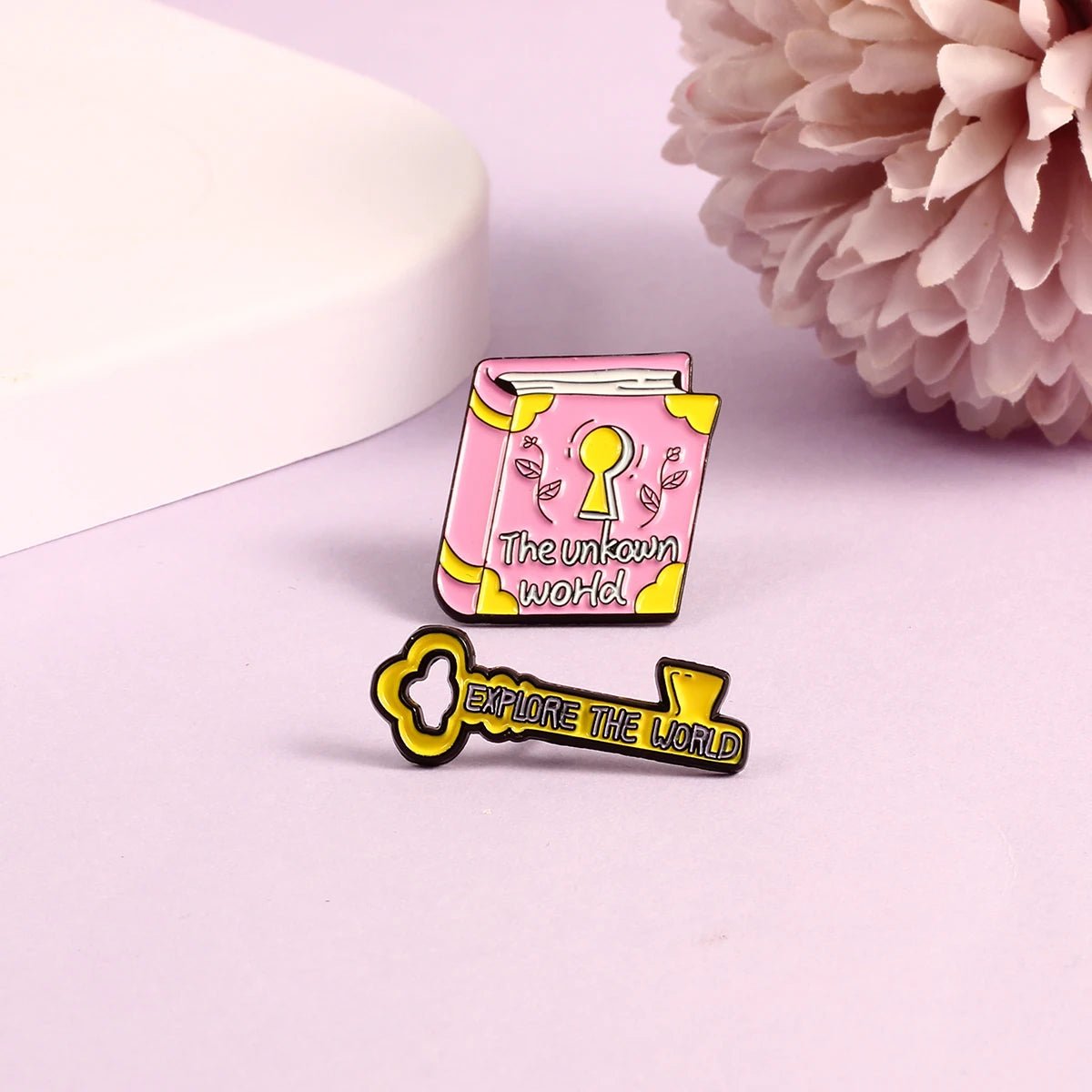 Pink book badge enamel pin with a keyhole and The unknown world text sits next to a yellow key enamel pin reading EXPLORE THE WORLD, displayed on a light purple surface with a pink flower.