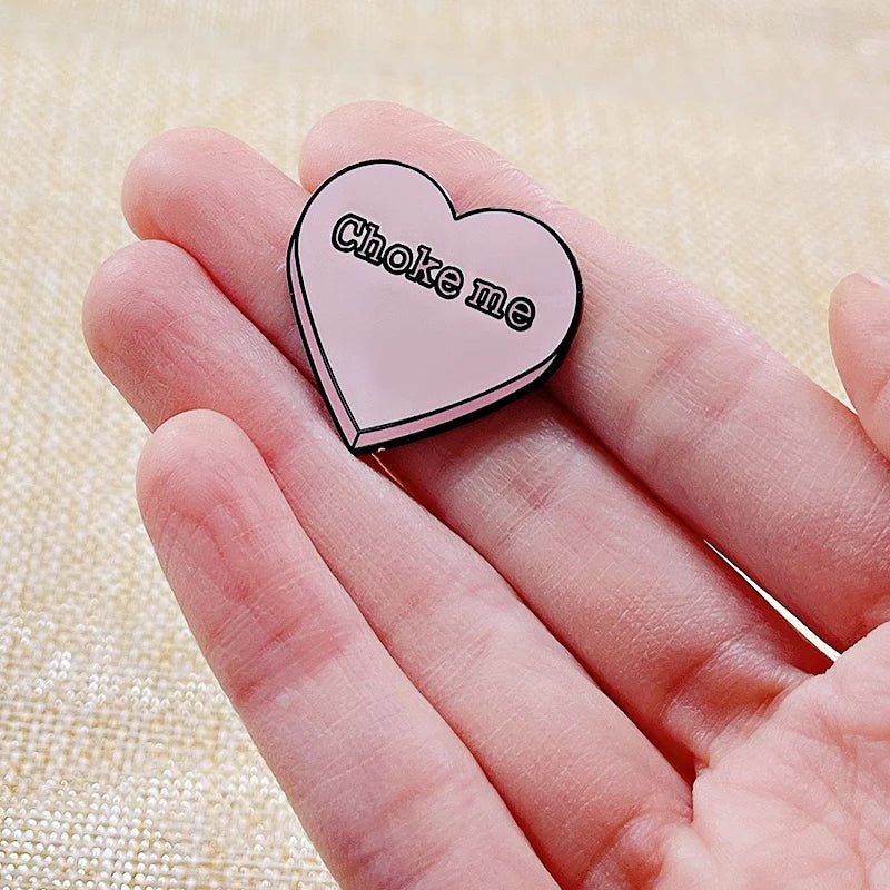 A hand holds a pink enamel pin by Kiss Me Later featuring the words Choke me in bold black text. The pin is shaped like a heart and stands out against a light, textured background.