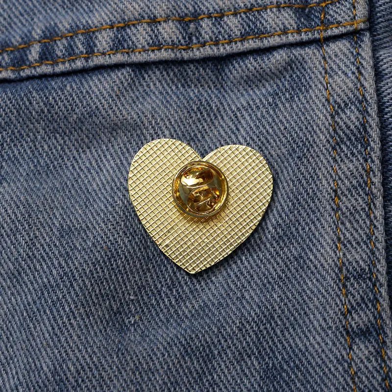 A Kiss Me Later gold textured love heart enamel pin with a clasp is attached to blue denim fabric with visible stitching.