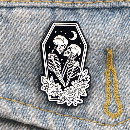 Black and white Skeleton Lovers enamel pin shows two skeletons embracing in a coffin-shaped frame, surrounded by flowers, with a crescent moon and stars in the background.