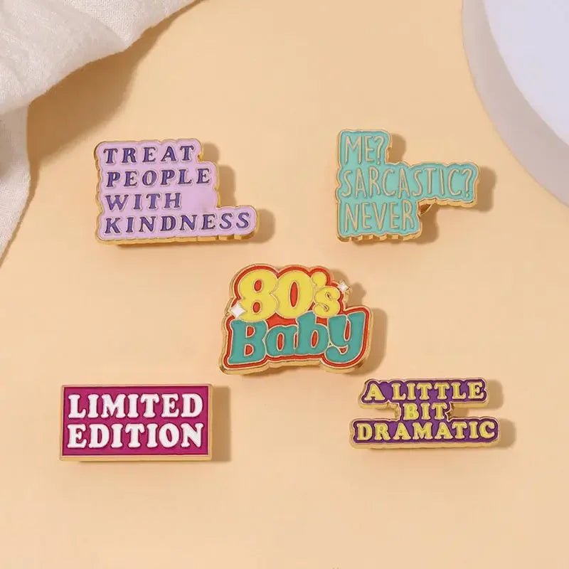 Set of five colorful enamel pins—including a red love heart—features phrases like TREAT PEOPLE WITH KINDNESS, ME? SARCASTIC? NEVER, 80s Baby, LIMITED EDITION, and A LITTLE BIT DRAMATIC on a beige background.