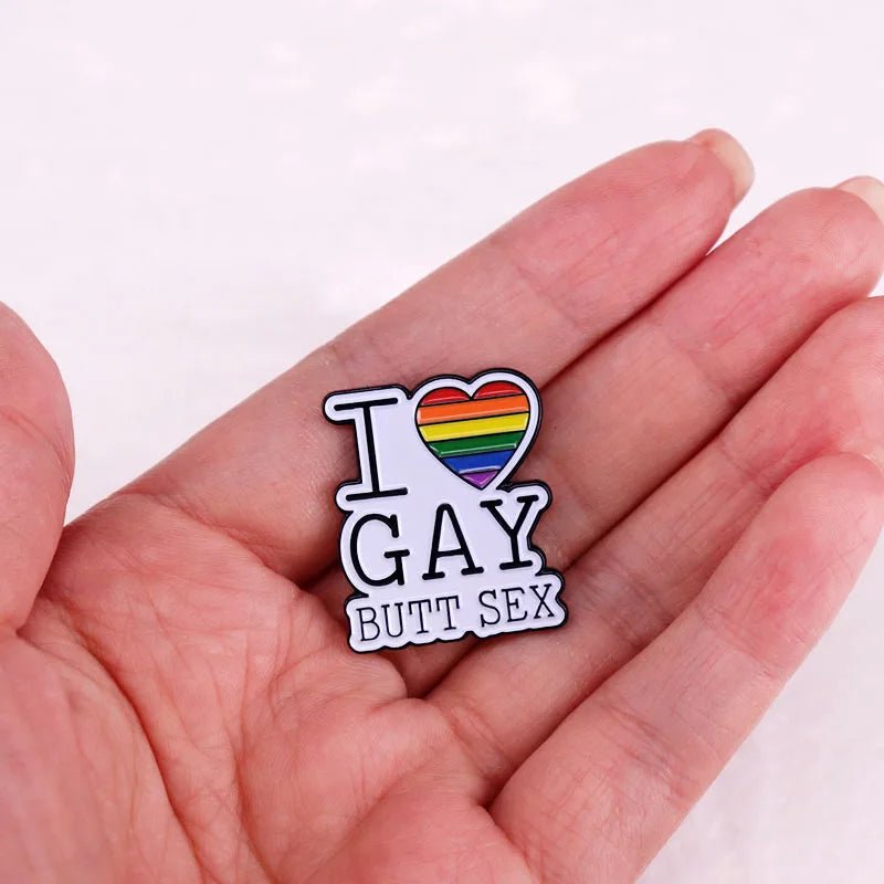 A hand holds a Kiss Me Later enamel pin shaped like a heart with rainbow stripes and the phrase I ♥ GAY BUTT SEX, celebrating LGBTQ+ pride.