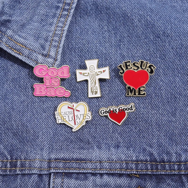 Five colorful Kiss Me Later enamel pins with messages like God is Bae, God is Good, and Jesus ♥ ME are pinned on blue denim. One is a cute pink cross, while a heart-shaped pin features an inspiring message inside.